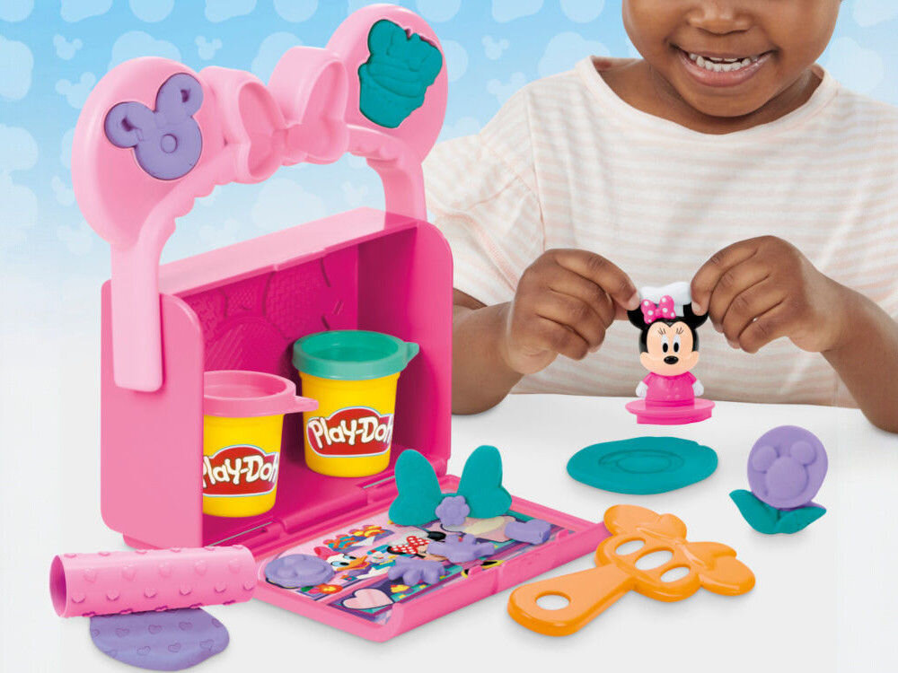 Ciastolina PLAY-DOH Disney Junior Mickey Mouse Przenośna cukiernia G31085X0 Dziecko bawiące się zestawem Play-Doh Disney Junior. Na stole znajduje się otwarta różowa walizeczka z foremkami, dwie tuby masy plastycznej, wałek w serduszka oraz wyciśnięte kształty z fioletowej i zielonej ciastoliny.