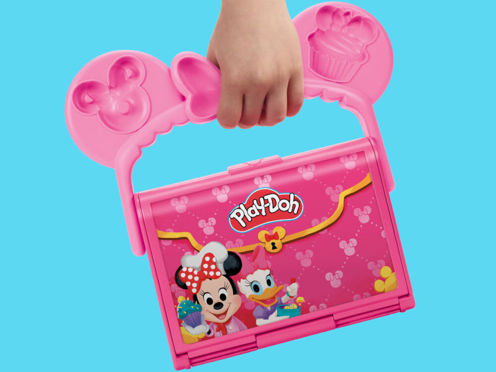 Ciastolina PLAY-DOH Disney Junior Mickey Mouse Przenośna cukiernia G31085X0 Ręka dziecka trzymająca zamkniętą różową walizeczkę Play-Doh z rączką w kształcie uszu Myszki Minnie. Na froncie grafika z Myszką Minnie i Kaczuszką Daisy piekącymi babeczki.