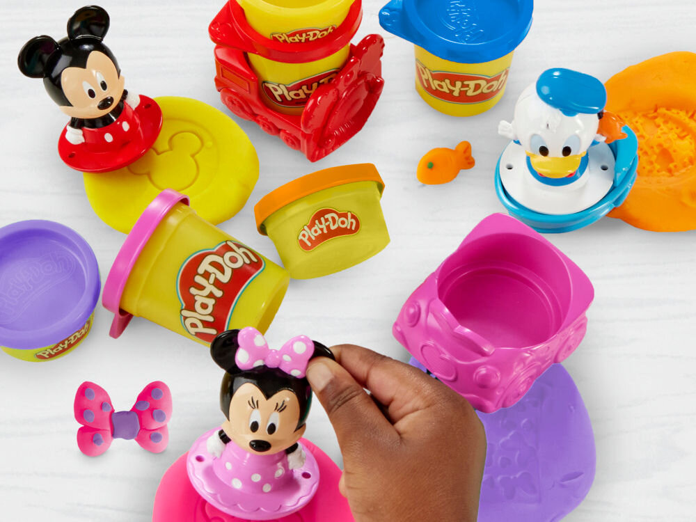 Ciastolina PLAY-DOH Disney Junior zestaw pieczątek G31105L0 Zbliżenie na dziecko bawiące się figurką Myszki Miki w stroju mechanika, która służy do odciskania pieczątek w żółtej ciastolinie Play-Doh. W tle widoczny fragment otwartej walizeczki z akcesoriami.