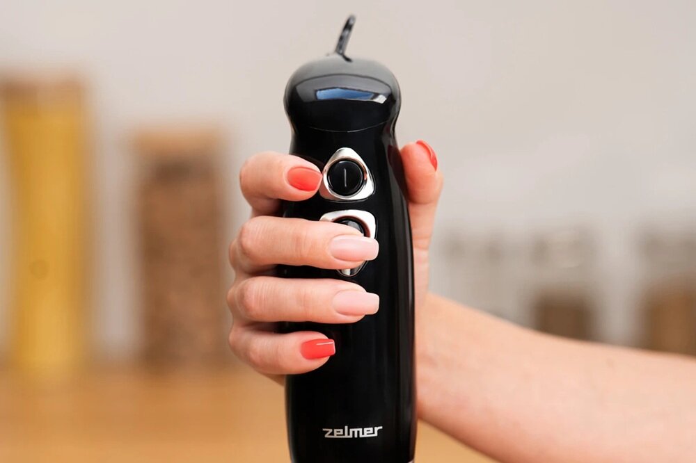 Osoba trzyma czarny blender ręczny z widocznym logo \'zelmer\'. Blender ma dwa przyciski na srebrnym panelu.