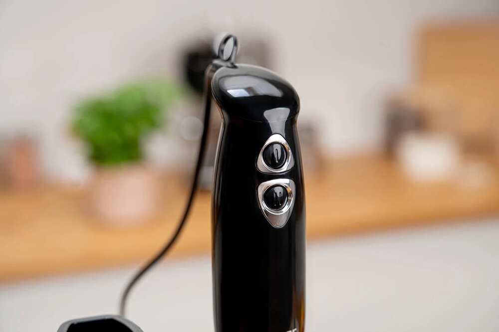 Czarny blender ręczny z dwoma przyciskami na uchwycie. W tle rozmyte elementy kuchenne, w tym zielona roślina w doniczce.