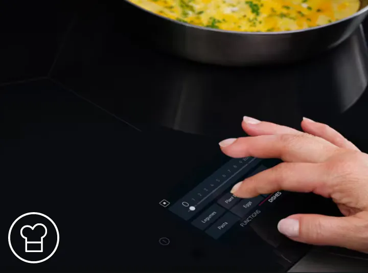 Osoba obsługuje panel dotykowy płyty indukcyjnej, na którym widoczne są opcje takie jak \'Lobster\', \'Pasta\', \'Eggs\', \'FUNCTIONS\', \'DISHES\'. W tle znajduje się patelnia z potrawą posypaną ziołami.