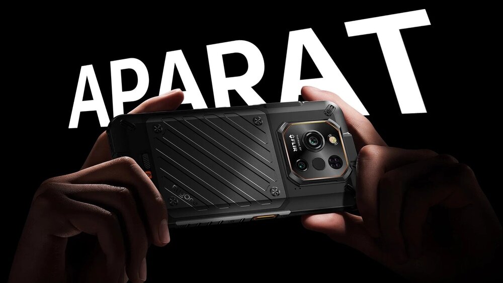 Osoba trzyma smartfon z widocznym aparatem fotograficznym na tylnej obudowie. W tle znajduje się duży, biały napis \'APARAT\'.