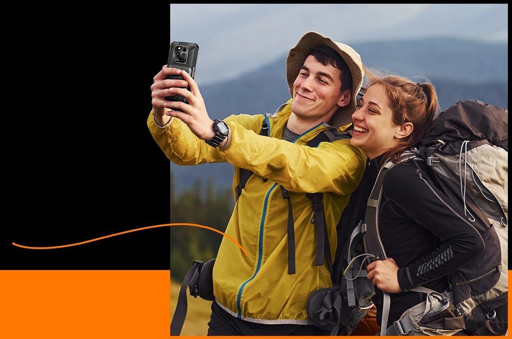 Dwie osoby w odzieży turystycznej robią sobie selfie smartfonem na tle górskiego krajobrazu. Osoba z lewej trzyma telefon w wyciągniętej ręce, a obie osoby są uśmiechnięte.