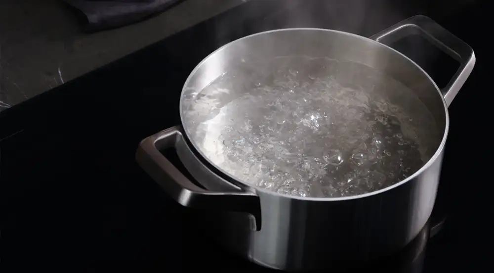 Na płycie indukcyjnej znajduje się garnek z gotującą się wodą. Para unosi się nad powierzchnią wody.