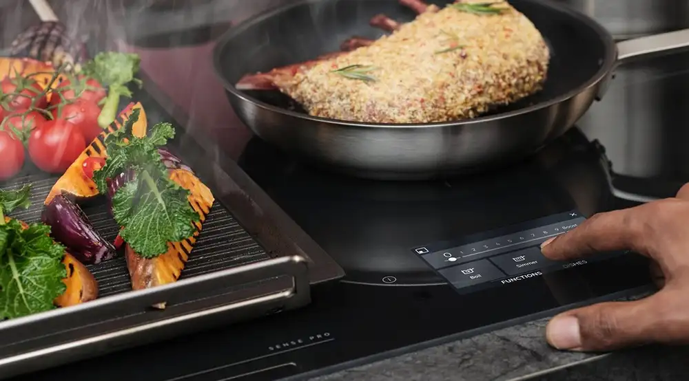 Na płycie indukcyjnej znajduje się patelnia z mięsem oraz grill z warzywami. Osoba dotyka panelu sterowania płyty, na którym widoczne są opcje \'Boil\', \'Simmer\' i \'Functions\'.