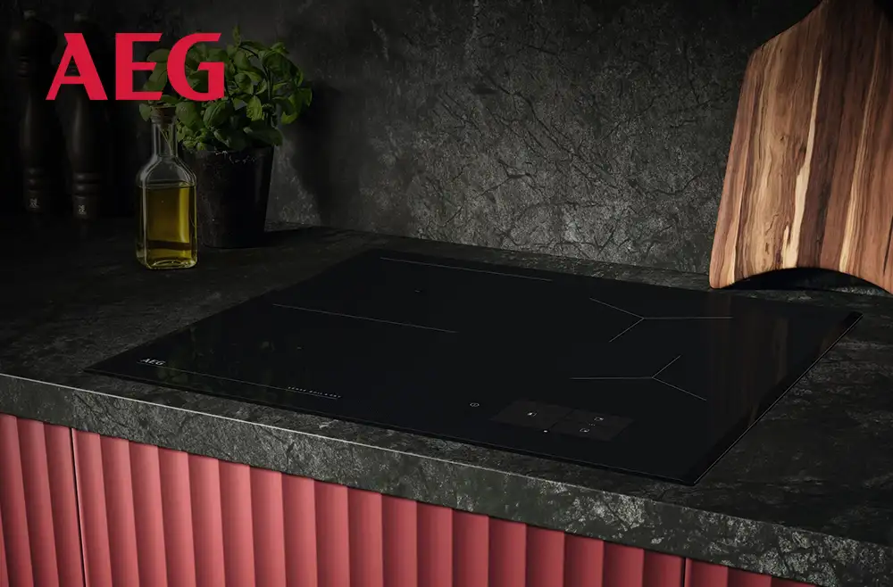 Czarna płyta indukcyjna wbudowana w ciemny blat kuchenny, obok której stoi butelka z oliwą i deska do krojenia. Na ścianie widoczne jest logo AEG.