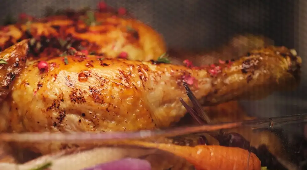 Zbliżenie na pieczonego kurczaka z przyprawami w szklanym naczyniu, otoczonego warzywami. Kurczak ma złocistą skórkę i jest udekorowany różowymi ziarnami pieprzu.