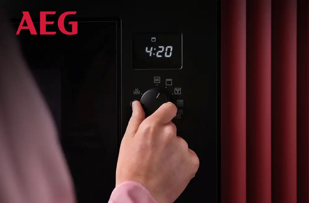 Osoba obsługuje czarną kuchenkę mikrofalową z wyświetlaczem pokazującym godzinę 4:20. Widoczne jest logo AEG oraz kilka ikon na panelu sterowania.