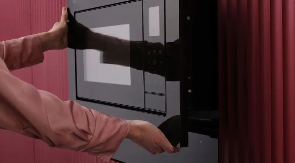 Osoba otwiera czarną kuchenkę mikrofalową wbudowaną w ścianę z czerwonymi panelami. Na panelu sterowania widoczne są przyciski dotykowe.