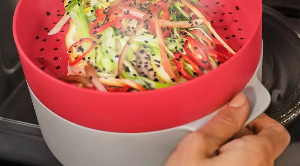 Osoba trzyma naczynie z czerwonym pokrywką, w którym znajdują się pokrojone warzywa posypane czarnym sezamem. Naczynie jest umieszczone w kuchence mikrofalowej.