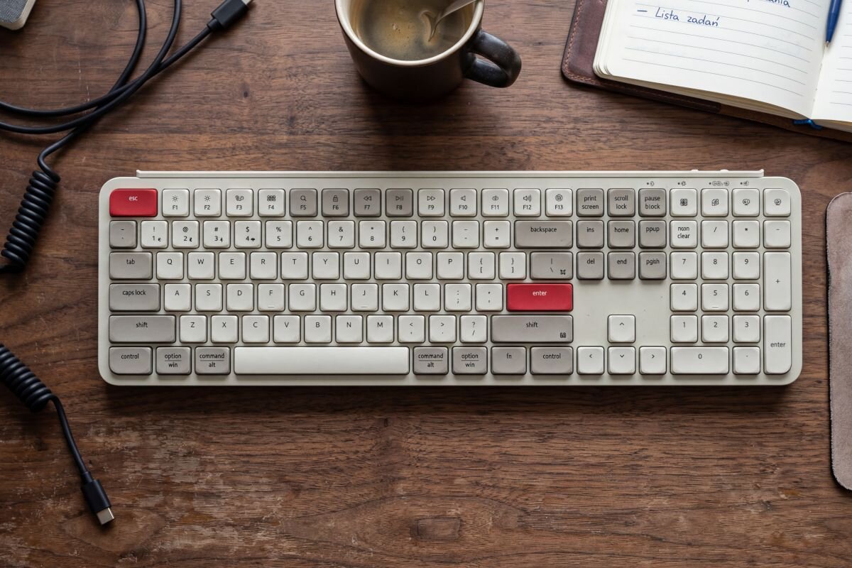 Keychron B6 Pro Keychron B6 Pro pełnowymiarowa, bezprzewodowa klawiatura lekka konstrukcja (623 g) trzy tryby łączności, Mechanizm nożycowy (scissor switch) pełne wsparcie dla ZMK Launcher - Klawiatura leżąca na drewnianym blacie roboczym w otoczeniu filiżanki kawy, notesu oraz przewodów zasilających.