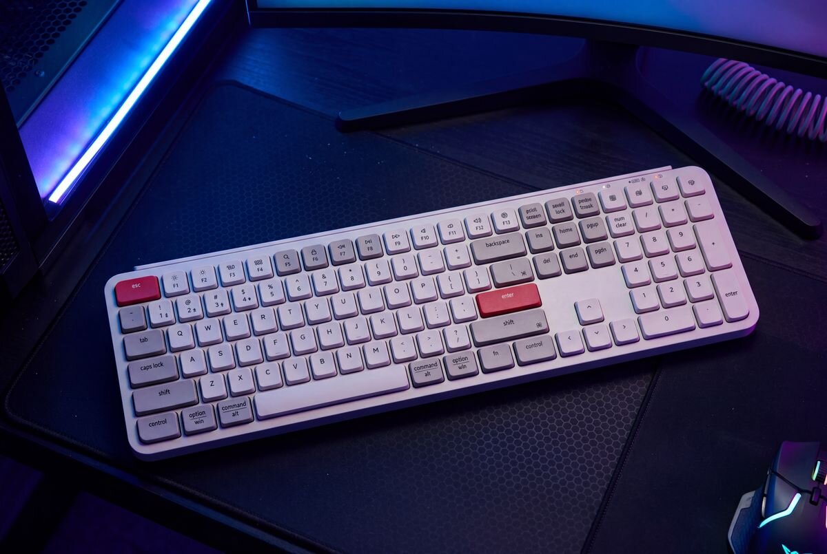 Keychron B6 Pro Keychron B6 Pro akumulator o pojemności 800 mAh 1200 godzin pracy na jednym ładowaniu 5 godzin dziennie 8 miesięcy działania pracę podczas ładowania przez USB-C - Klawiatura umieszczona na stanowisku komputerowym z widocznym fragmentem monitora oraz podświetleniem gamingowym.