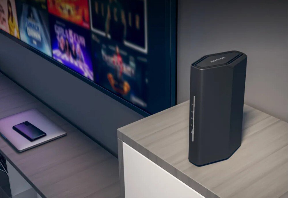 Router NETGEAR Nighthawk RS100 Pionowy router o sześciokątnej podstawie stoi na szafce obok włączonego telewizora, na którym widać kafelki z filmami, opis