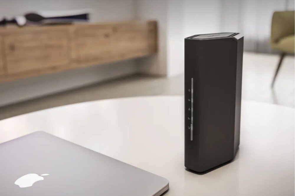 Router NETGEAR Nighthawk RS100 Router stoi na jasnym stole w salonie, w tle widać rozmyty fragment drewnianej komody, a na pierwszym planie zamknięty laptop, zabezpieczenia, firewall, szyfrowanie