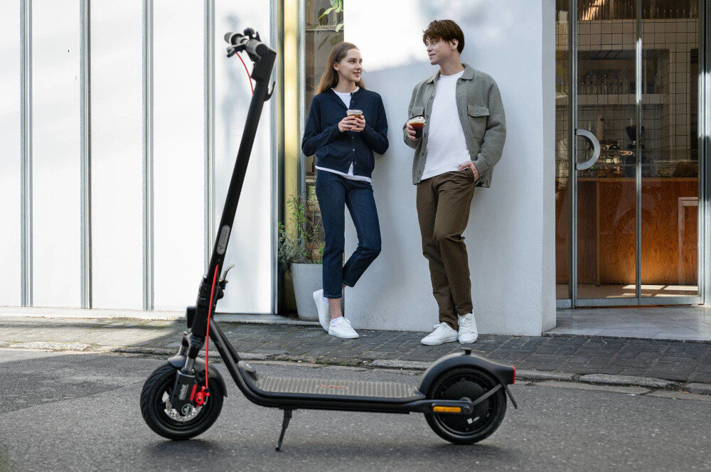 Hulajnoga elektryczna SEGWAY F2 Pro D II 65km 900W 10 cali Czarny miasto chodnik kawiarnia dwie osoby rozmawiają ze sobą oparte o ścianę na pierwszym planie stoi hulajnoga oparta o stopkę składana stopka nóżka zamontowana w centralnym punkcie ciężkości głośny elektryczny klakson bezpieczeństwo