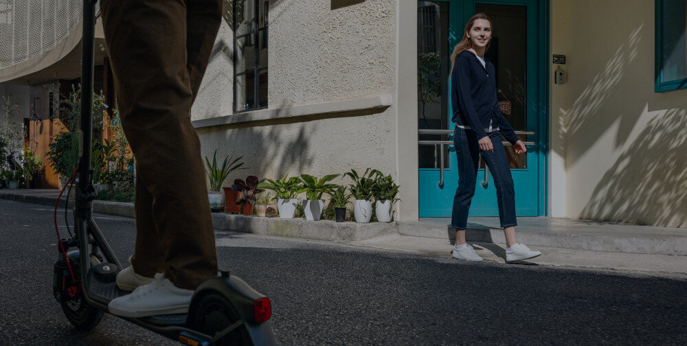 Hulajnoga elektryczna SEGWAY F2 Pro D II 65km 900W 10 cali Czarny miasto osoba idzie po chodniku patrzy na drugą osobę jadącą na hulajnodze w tle budynek bateria o pojemności 12,8 Ah 460 Wh zasięg do 40 km w trybie ECO w trybie SPORT do 50 km bateria litowo-jonowa napięcie 36 V czas ładowania około 8 godzin