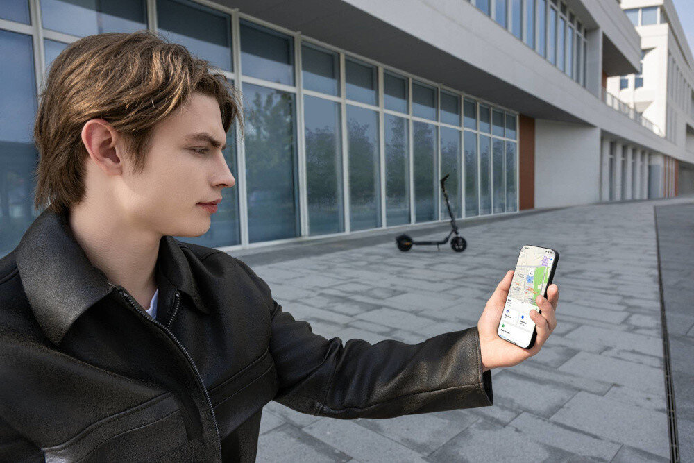 Hulajnoga elektryczna SEGWAY F2 Pro D II 65km 900W 10 cali Czarny miasto budynek osoba trzyma w dłoni telefon z włączoną aplikacją w tle stoi hulajnoga wbudowany moduł Bluetooth aplikacja mobilna Segway-Ninebot Smart App na systemy Android i iOS