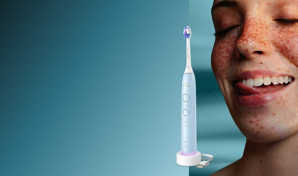 Zbliżenie na twarz uśmiechniętej kobiety, która językiem przesuwa po zębach. Obok szczoteczka soniczna.