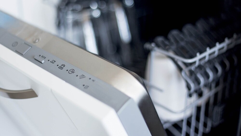 Zbliżenie na panel sterowania zmywarki z widocznymi przyciskami i ikonami. W tle widać wnętrze zmywarki z koszem na naczynia.
