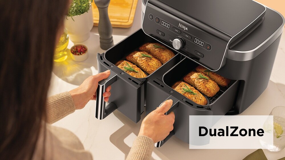 Osoba otwiera podwójny kosz frytkownicy beztłuszczowej marki NINJA, w którym znajdują się pieczone potrawy. Na obrazie widoczny jest napis \'DualZone\'.