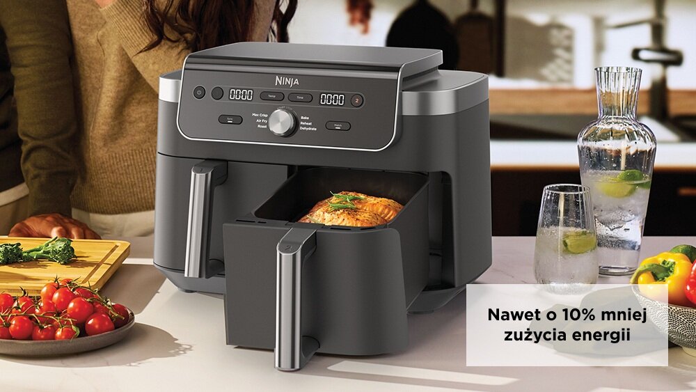 Na blacie kuchennym stoi frytkownica beztłuszczowa z podwójnym koszem, w jednym z koszy znajduje się pieczony kurczak. Obok urządzenia leży deska z pomidorami, a na blacie stoi szklanka z napojem i karafka z wodą; widoczny jest tekst: \'Nawet o 10% mniej zużycia energii\'.