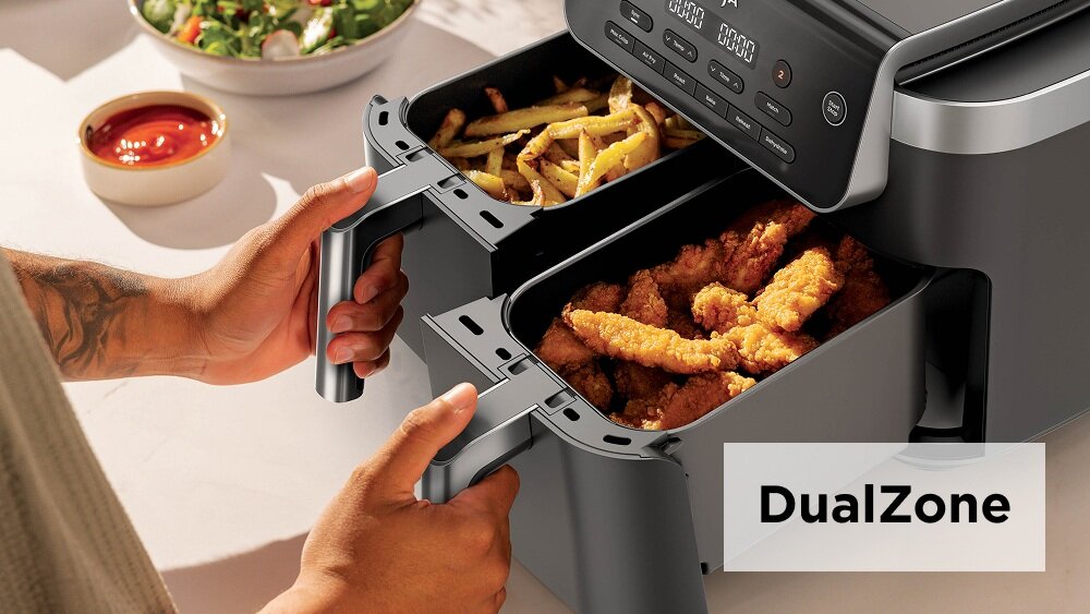 Osoba trzyma frytkownicę beztłuszczową z podwójnym koszem, w którym znajdują się frytki i kawałki kurczaka. Na obrazie widoczny jest napis \'DualZone\'.
