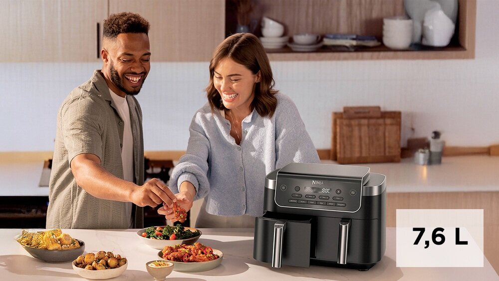 Na blacie kuchennym stoi frytkownica beztłuszczowa z podwójnym koszem marki Ninja. Obok niej znajdują się miski z jedzeniem, a w tle widać kuchenne półki z naczyniami.