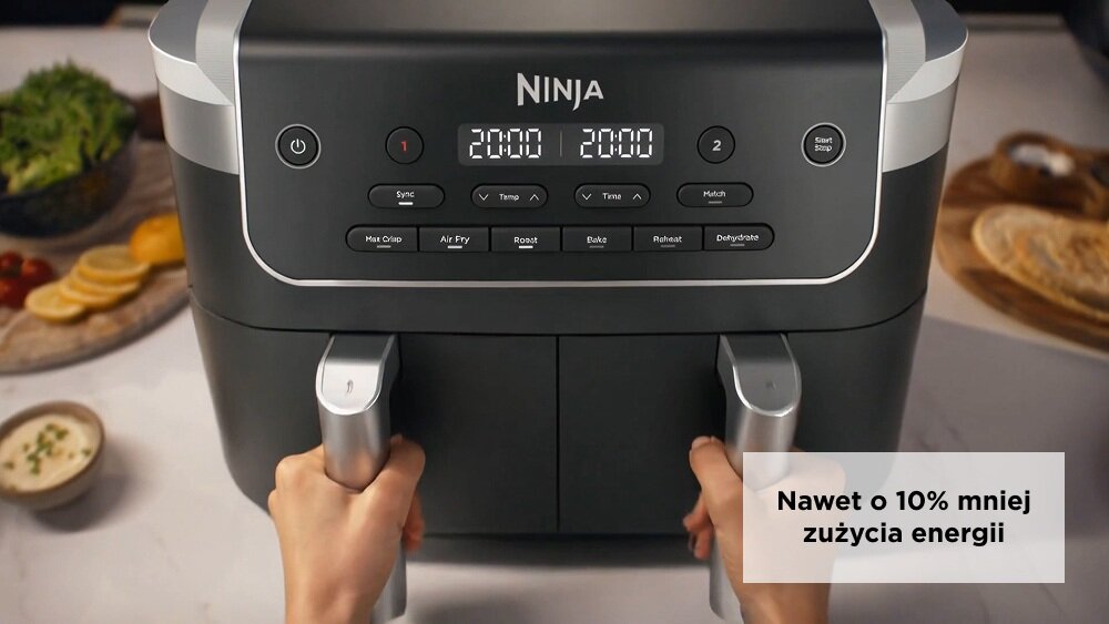 Na obrazie widoczna jest frytkownica beztłuszczowa z logo \'Ninja\' na panelu sterowania, z wyświetlaczami pokazującymi \'2000\'. Na dole widoczne są dwie ręce trzymające uchwyty koszy, a w prawym dolnym rogu znajduje się napis \'Nawet o 10% mniej zużycia energii\'.