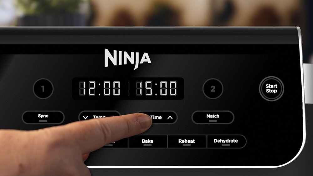 Panel sterowania frytkownicy beztłuszczowej z logo \'NINJA\' na górze, wyświetlający czas 12:00 i 15:00. Widoczny jest palec naciskający przycisk \'Time\', a poniżej znajdują się przyciski \'Bake\', \'Reheat\' i \'Dehydrate\'.