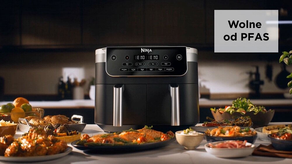 Na blacie kuchennym stoi frytkownica beztłuszczowa z podwójnym koszem marki Ninja. Obok niej znajdują się różnorodne potrawy, a w rogu widnieje napis \'Wolne od PFAS\'.