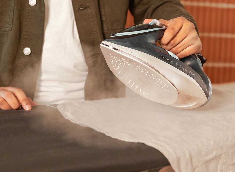Osoba prasuje tkaninę przy użyciu żelazka parowego. Na żelazku widoczne jest logo \'cecotec\' oraz napis \'IronHero 2600\'.