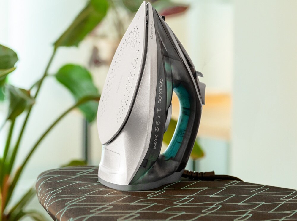 Na zdjęciu znajduje się żelazko o srebrnej stopie, ustawione na desce do prasowania. Na obudowie widoczny jest napis \'cecotec\' oraz oznaczenie \'2600W\'.