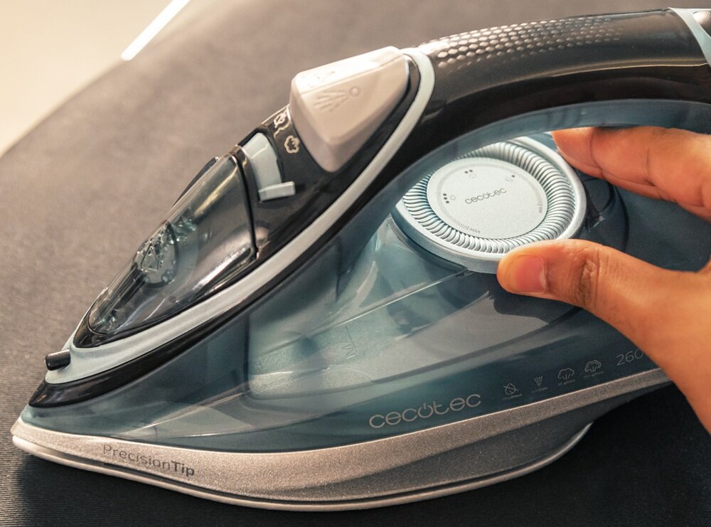 Zdjęcie przedstawia żelazko z widocznym logo \'cecotec\' na przezroczystej obudowie. Osoba trzyma pokrętło regulacji temperatury.