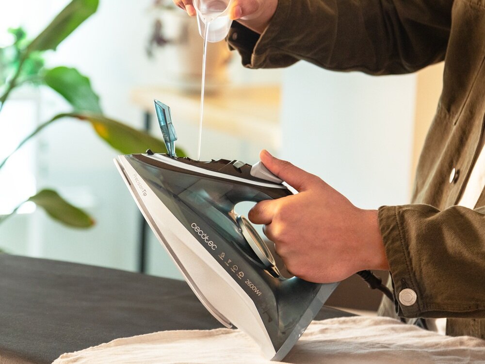 Osoba nalewa wodę do żelazka marki Cecotec, model IronHero 2600. W tle widoczna jest roślina doniczkowa.