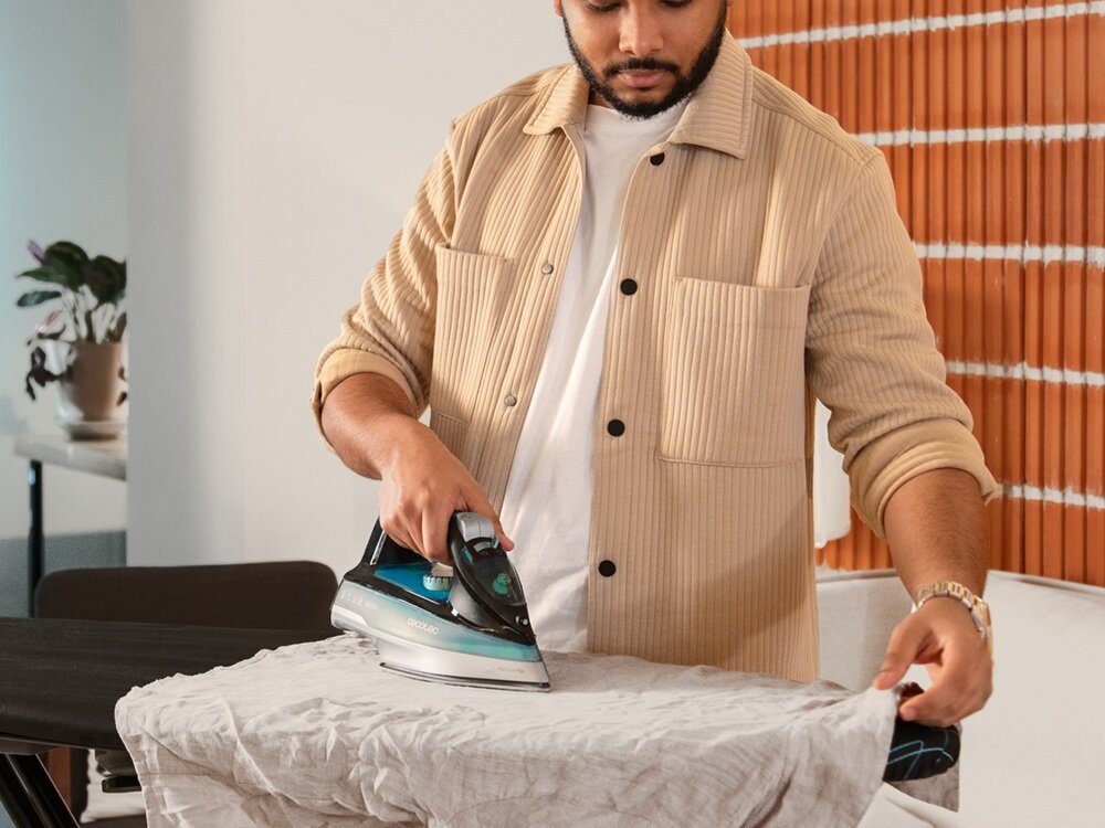 Osoba prasuje tkaninę przy użyciu żelazka z widocznym napisem \'CECOTEC\'. W tle znajduje się pomieszczenie z rośliną w doniczce.