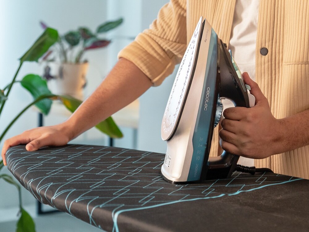 Osoba trzyma żelazko CECOTEC IronHero 2600 Force Steam na desce do prasowania z geometrycznym wzorem. W tle widoczne są rośliny doniczkowe.