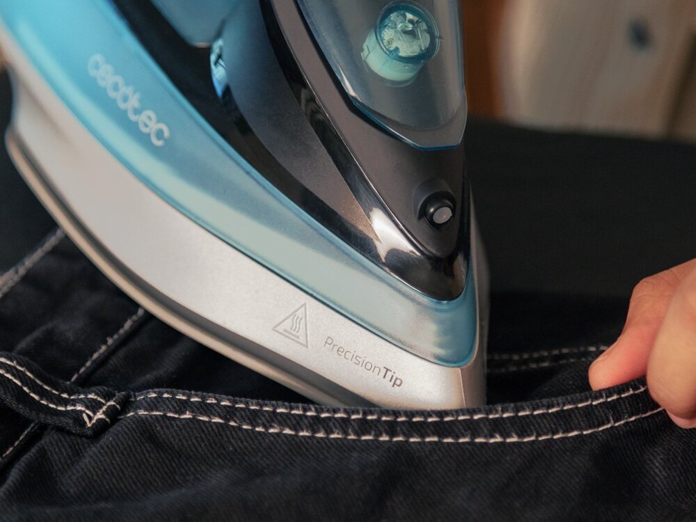 Żelazko z niebieskimi i srebrnymi elementami prasuje ciemny materiał z białymi szwami. Widoczny jest napis \'cecotec\' oraz \'PrecisionTip\'.