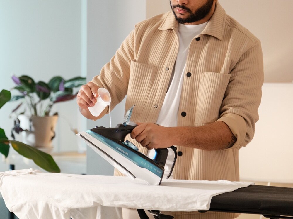 Osoba nalewa wodę do żelazka marki CECOTEC, które znajduje się na desce do prasowania. W tle widoczne są rośliny doniczkowe i jasne wnętrze.