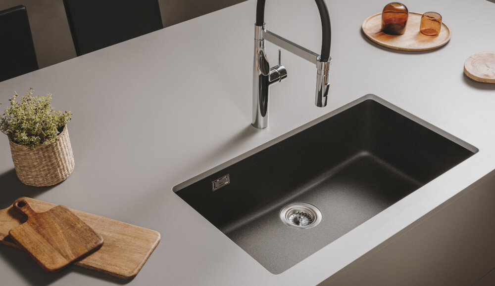 Na obrazie widoczny jest czarny zlewozmywak jednokomorowy wbudowany w jasny blat kuchenny. Obok znajdują się drewniane deski do krojenia i roślina w koszyku.