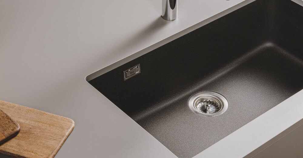 Czarny zlewozmywak jednokomorowy z metalowym odpływem i chromowaną baterią. Obok znajduje się drewniana deska do krojenia.