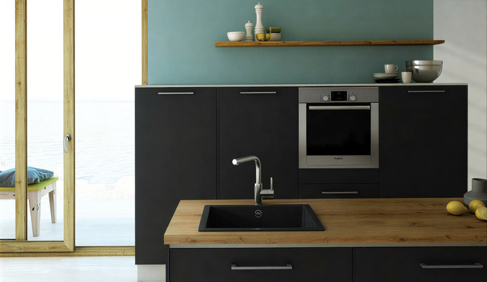 Kuchnia z czarnymi szafkami i drewnianym blatem, na którym znajduje się czarny zlewozmywak jednokomorowy z kranem. W tle widać piekarnik w zabudowie oraz półkę z dekoracjami.
