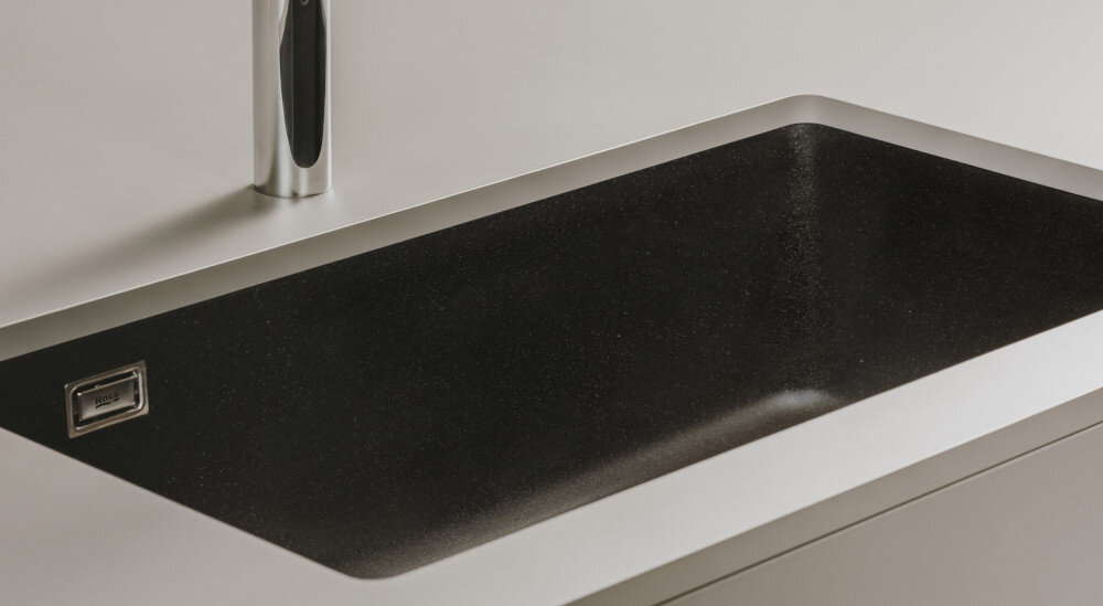 Na obrazie widoczny jest czarny zlewozmywak jednokomorowy wbudowany w jasny blat. Obok zlewozmywaka znajduje się chromowana bateria kuchenna.