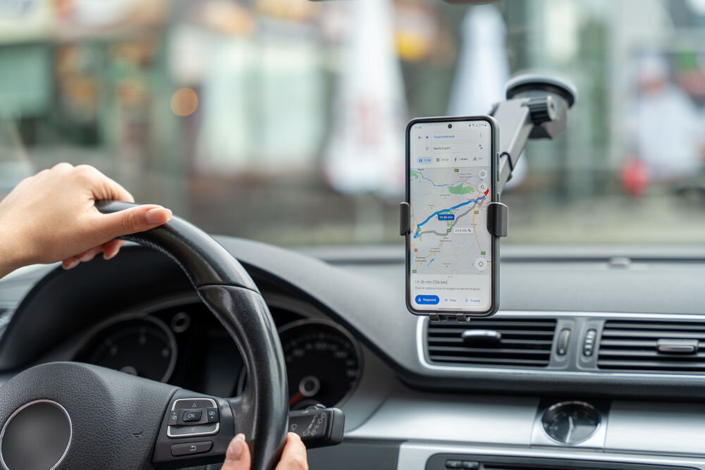 Wnętrze samochodu z zamontowanym na szybie uchwytem na telefon, w którym znajduje się smartfon z włączoną aplikacją map. Osoba trzyma kierownicę, a na ekranie telefonu widoczna jest trasa na mapie.