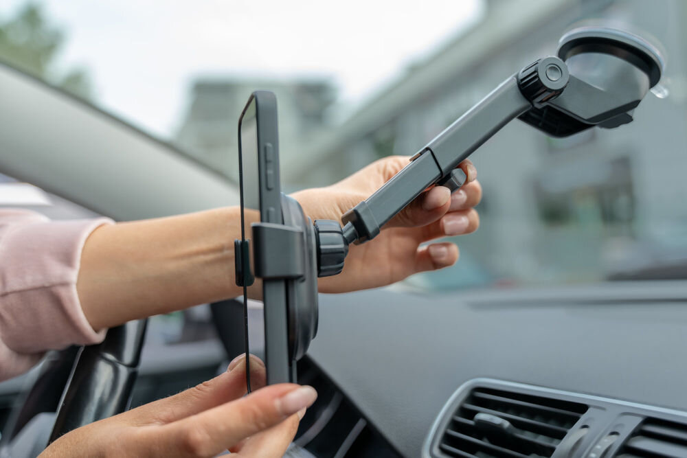 Osoba montuje czarny uchwyt samochodowy na szybę, trzymając w nim smartfon. W tle widoczna jest deska rozdzielcza samochodu.