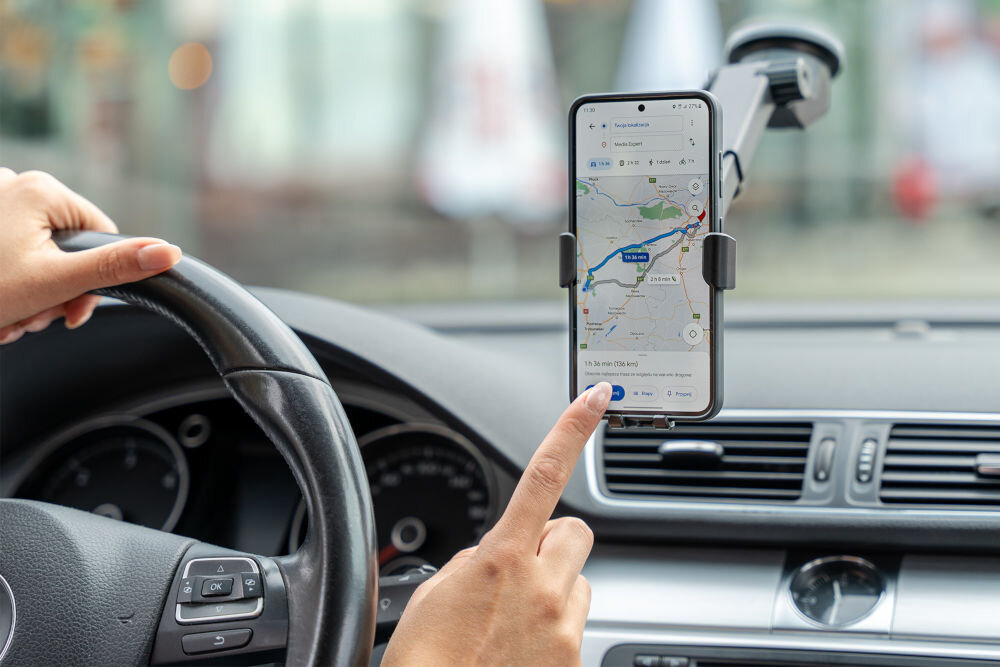W samochodzie zamontowany jest czarny uchwyt na telefon, w którym znajduje się smartfon z włączoną aplikacją map. Osoba trzyma kierownicę i dotyka ekranu telefonu.