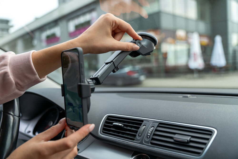 Osoba montuje czarny uchwyt samochodowy na szybę, w którym znajduje się smartfon. W tle widoczne są budynki i parasole na zewnątrz.