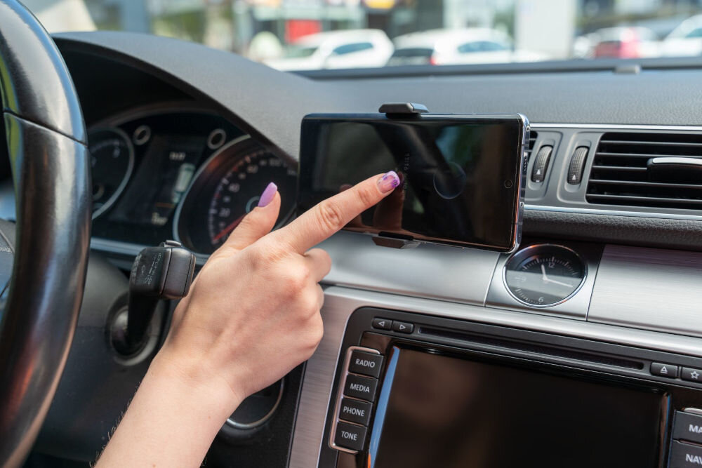 Uchwyt samochodowy WANDOO WCH100 stabilne mocowanie szczęki telefon nawigacja bezpieczna jazda ręce na kierownicy Dłoń z fioletowym manicure dotyka ekranu smartfona zamontowanego na desce rozdzielczej samochodu.