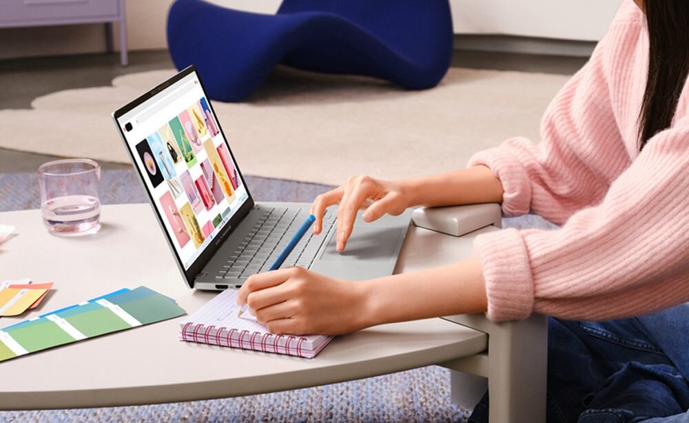 Osoba siedzi przy stole, używając laptopa z otwartą stroną internetową z kolorowymi grafikami. Obok laptopa leży notatnik i próbki kolorów, a na stole stoi szklanka z wodą.
