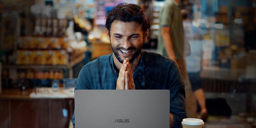 Mężczyzna pracuje na laptopie ASUS VivoBook 16 w kawiarni, skupiając się nad ekranem urządzenia – Komfort i funkcje użytkowe